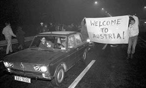 Zu sehen ist ein Auto mit zwei Personen daneben. Die zwei Leute halten ein Plakat mit der Aufschrift "WELCOME TO AUSTRIA!".